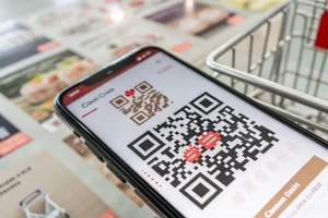Close-up view of a smartphone screen showing a large, eye-catching QR code, surrounded by faint e-commerce elements like shopping carts. Bright lighting emphasizes the QR code's design against a soft background.