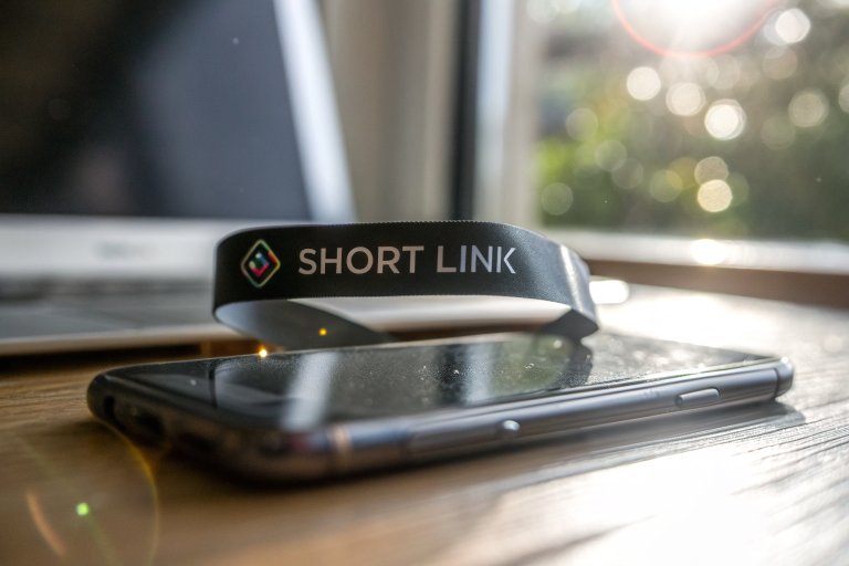 A close-up of a holographic short URL ribbon, displaying a branded link above a smartphone, with a blurred email compose window in the background. The image emphasizes trust and clickability with modern typography and soft lighting effects.